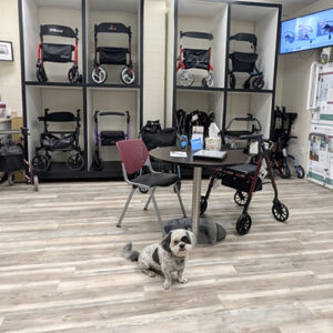 Multiple Walkers and Rollators on a shelf in our fall prevention room. A table with a chair and a rollator is in the middle of the room with a dog sitting in front of them.