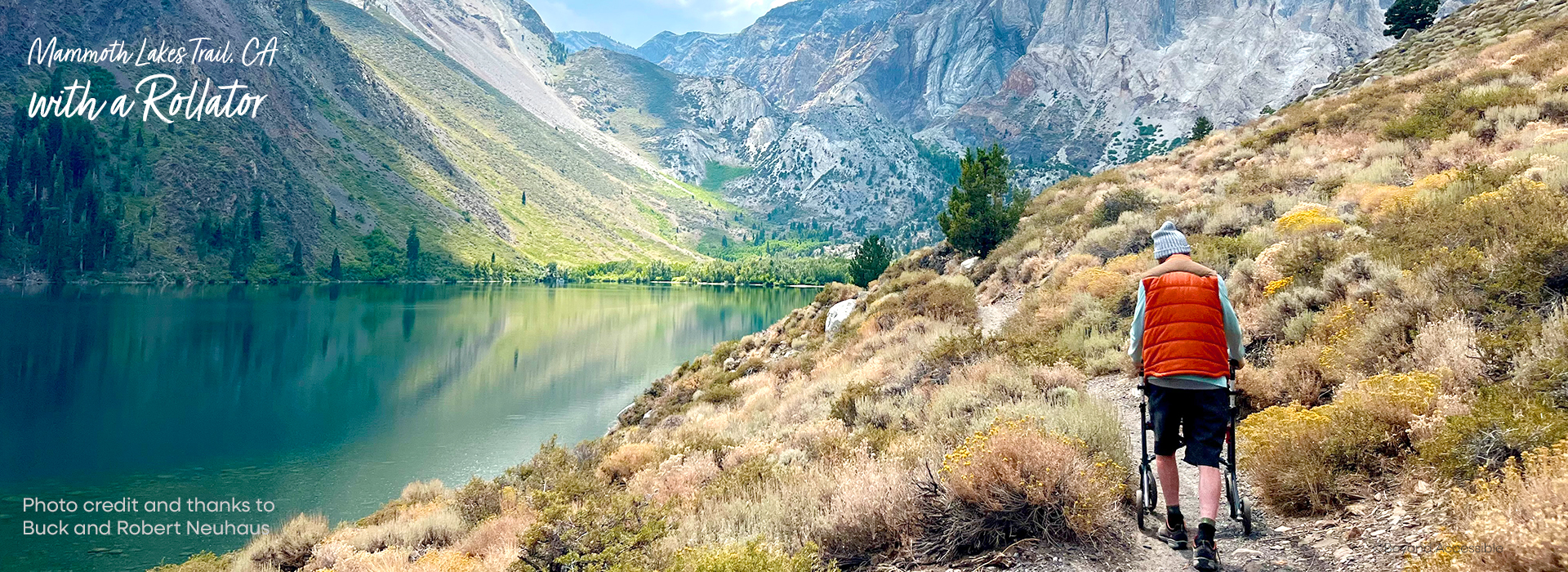 photo of a man wearing a red vest and black shorts pushing a rollator on a path next to a scenic lake and mountain side.