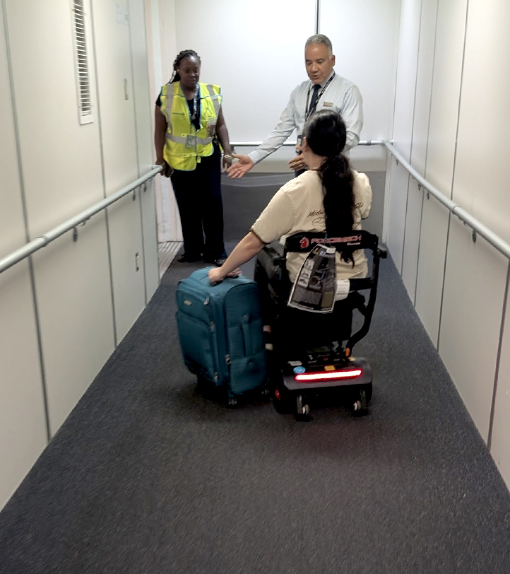 woman on a scooter, guiding a suitcase next to her talking to two people on the walkway
